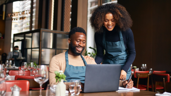 A restaurant manager in an apron looks at a laptop while seated in the dining area of the restaurant, while another worker looks over his shoulder.