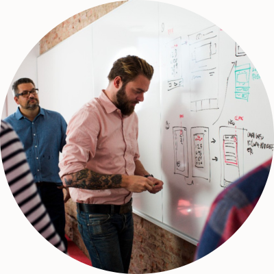 A man with his shirtsleeves rolled up looks at a technical drawing on a whiteboard while colleagues look on.