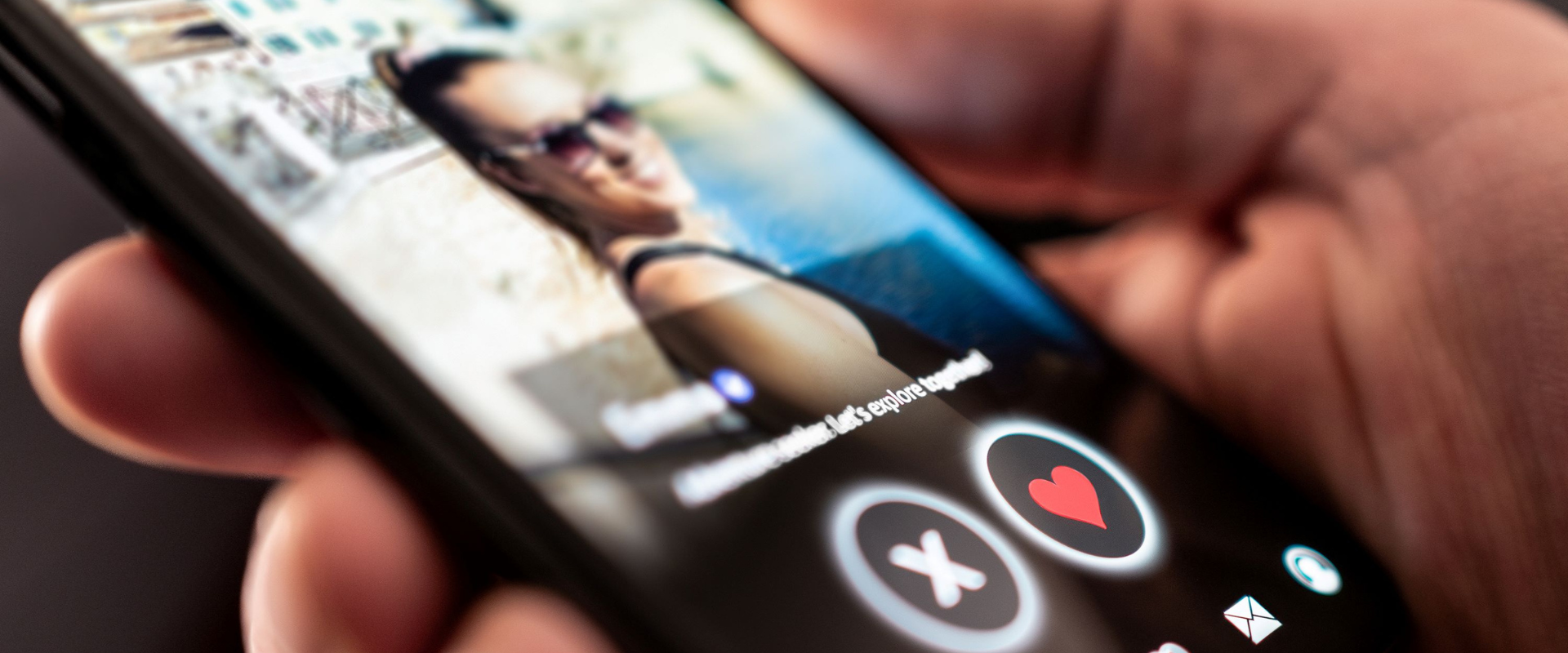 A hand holding a phone with a woman's photo on a dating app and buttons for X and a heart. 