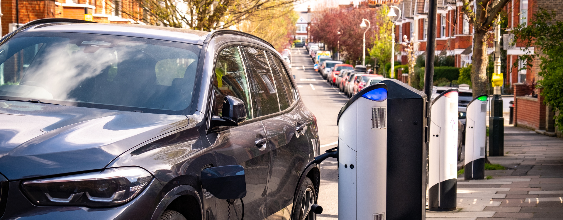 An EV sedan at a curb in a city plugged into an EV charging device. 