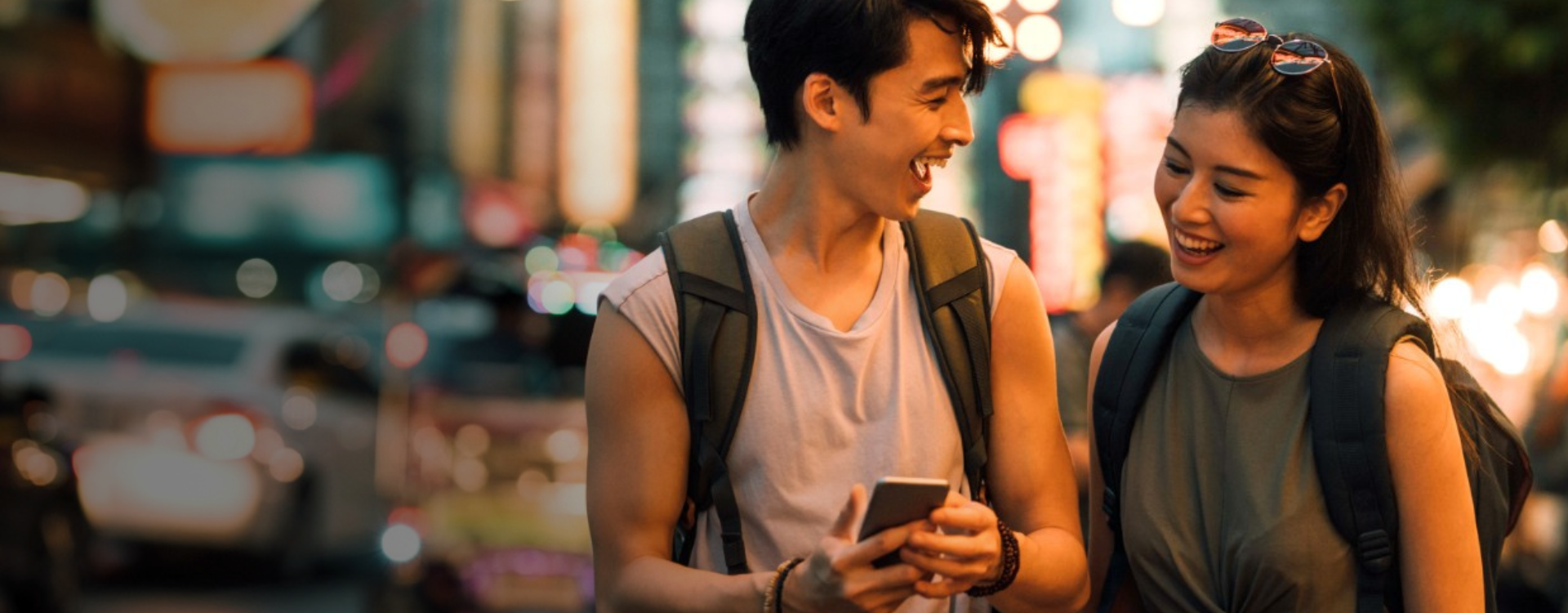 Two young people walk along a busy city street, with the man holding a cell phone and showing it to his female companion. 