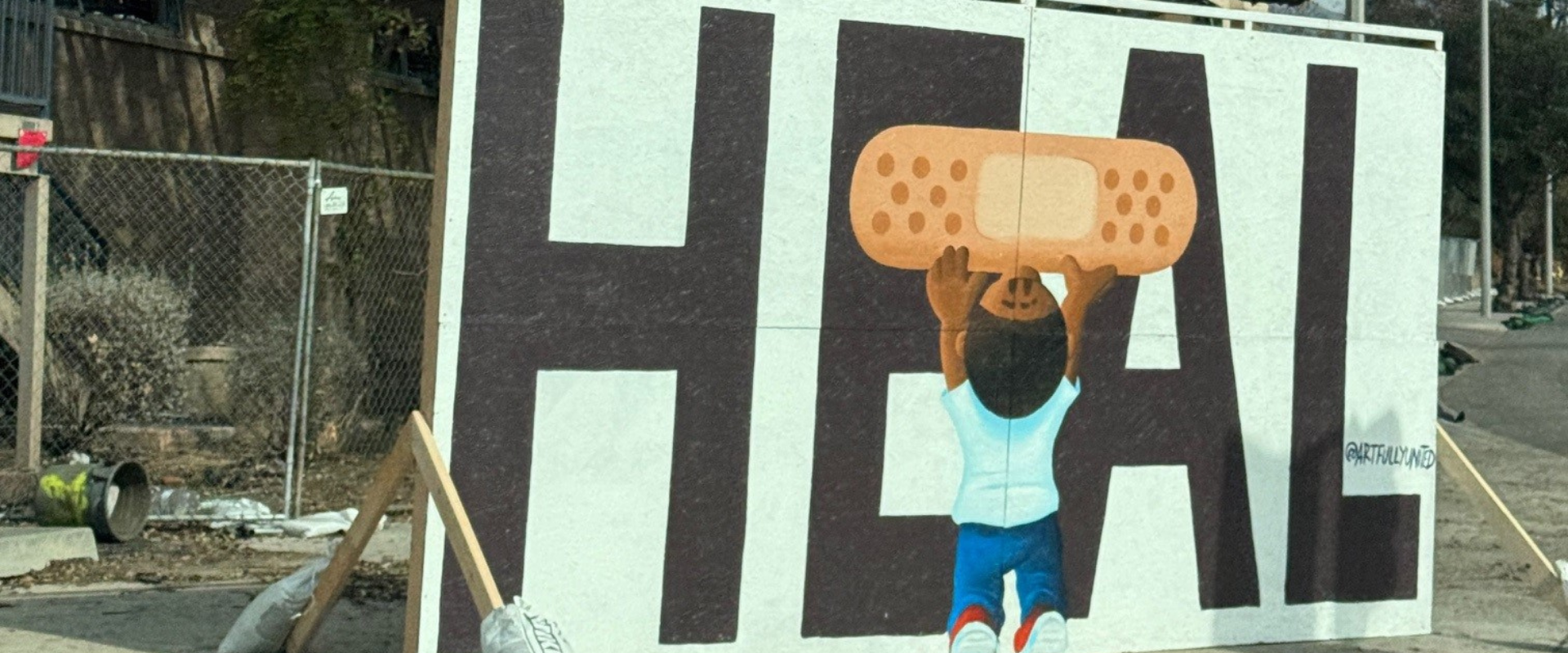 A sign saying "HEAL" with a small child holding a Band-Aid over the E and A, in front of wildfire destruction.