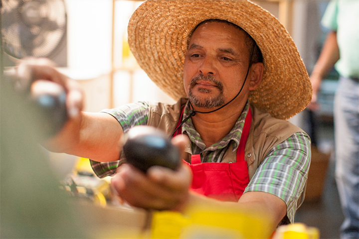 A man in a straw hat and aprons arranges vegetables at a farm stand. 