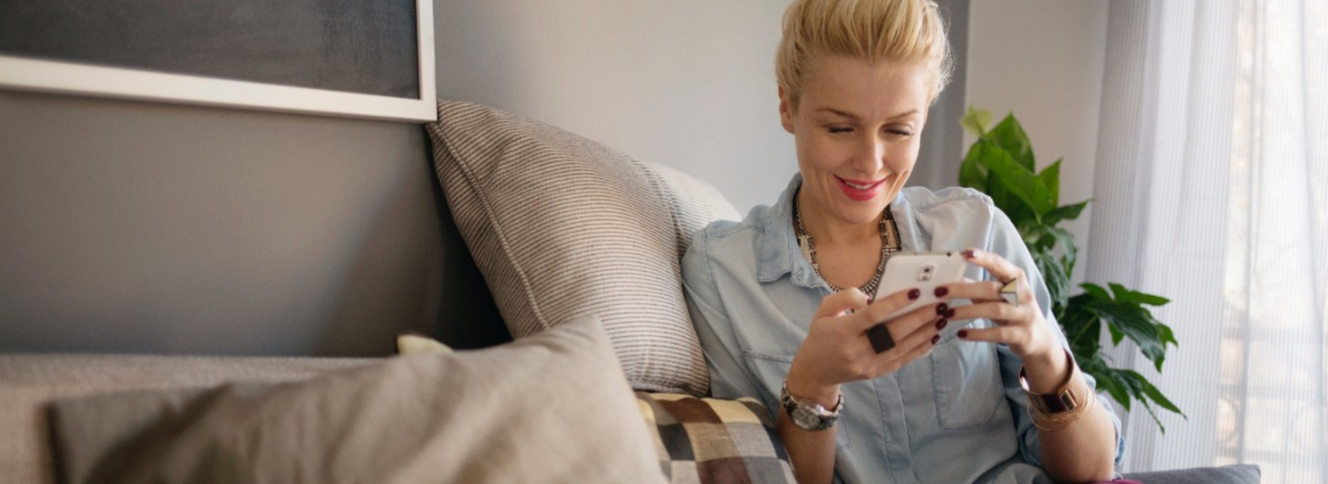 A woman smiles at her phone while seated on a couch in her home.
