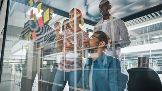 Five people gather in front of a glass wall in the office examining sticky notes in a brainstorming session.