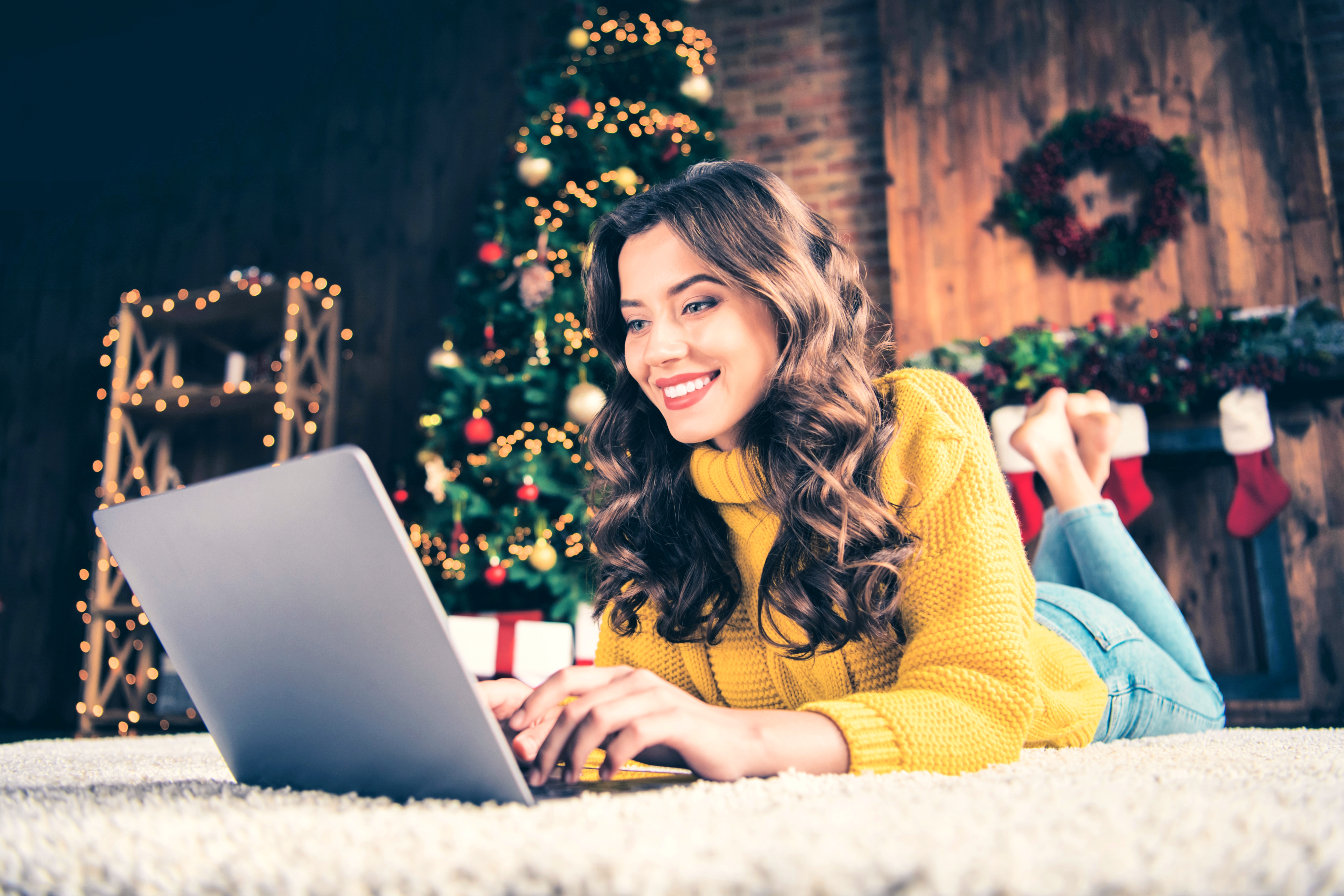 A woman in a yellow sweater lies on the floor in front of a Christmas tree looking at her laptop. 