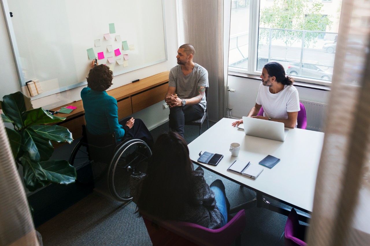 A woman in a wheelchair points to a whiteboard while three colleagues seated in chairs look on. 