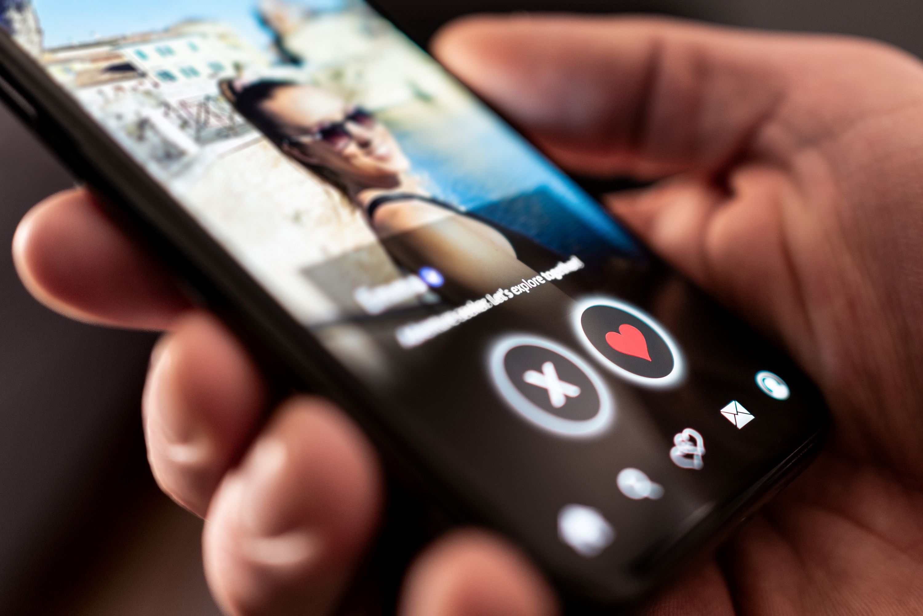 A hand holding a phone with a woman's photo on a dating app and buttons for X and a heart. 