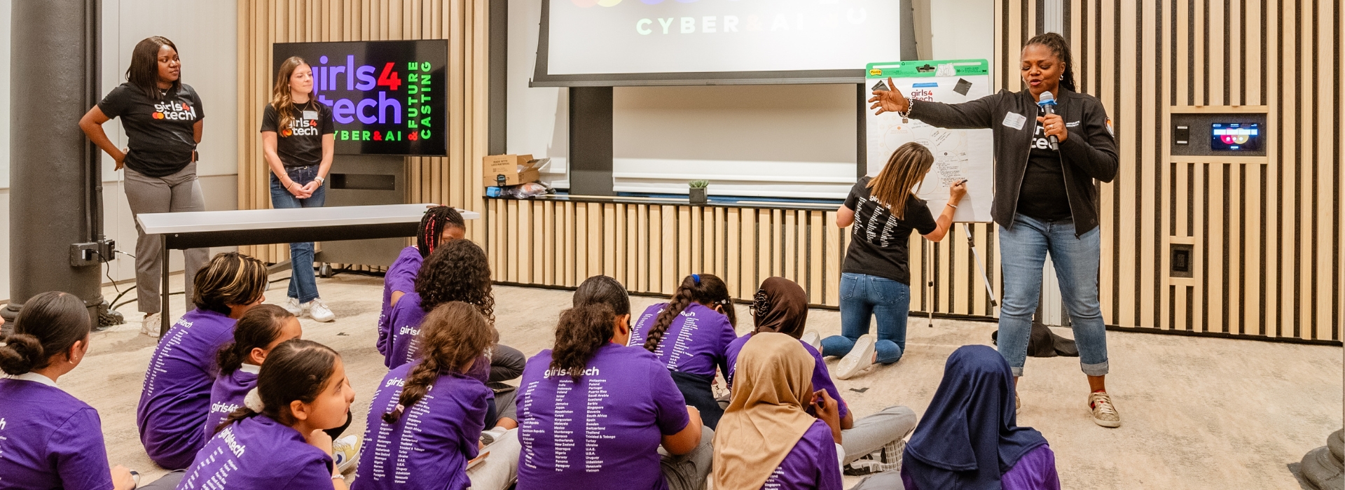 Alissa "Dr. Jay" Abdullah stands in front of a group of young female students in purple Girls4Tech T-shirts, seated on the floor, to discuss future cyber risks.