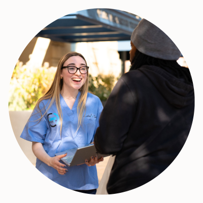 A woman in hospital scrubs holding a laptop talks to another woman.