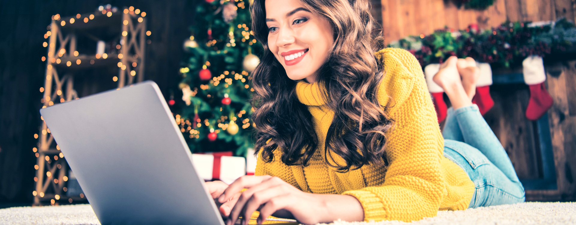 A woman in a yellow sweater lies on the floor in front of a Christmas tree looking at her laptop. 