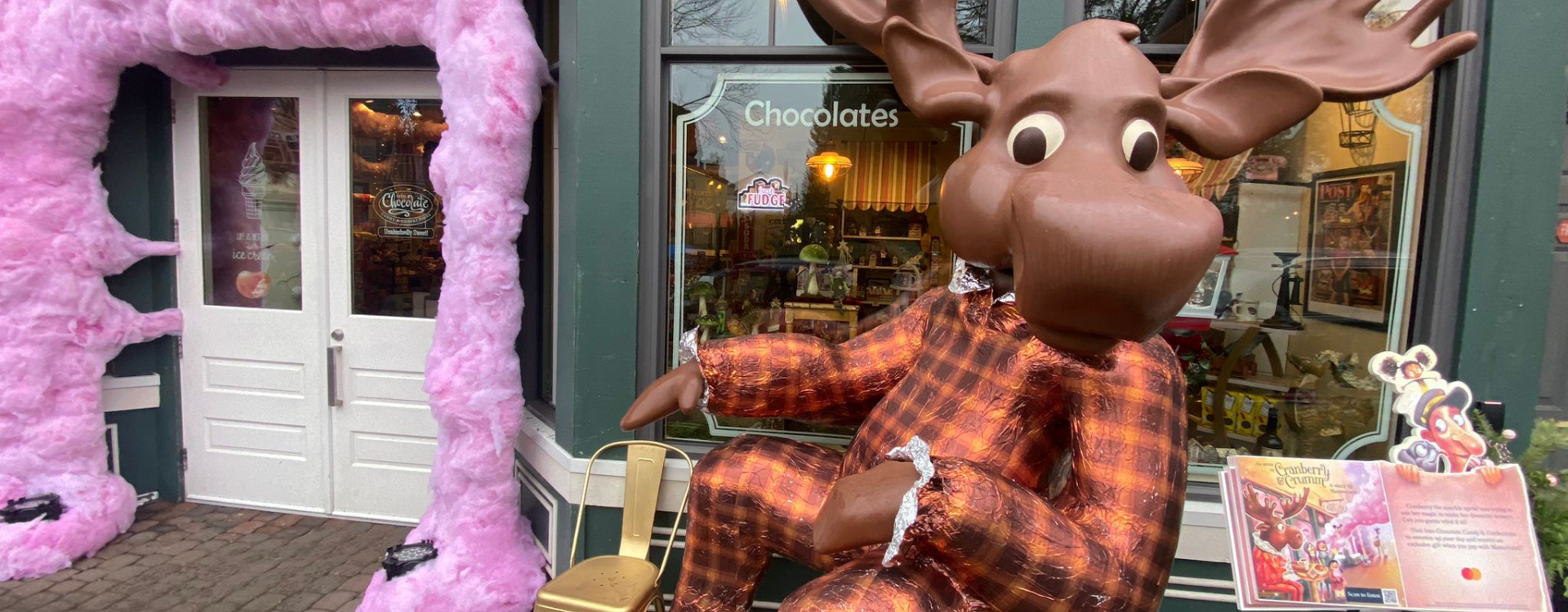 An anthropomorphic moose dressed in a suit sits in front of a chocolate shop next to Mastercard's holiday-themed storybook.